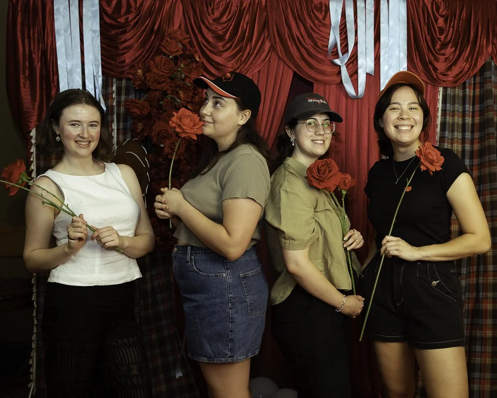Four young women smiling and holding red roses, standing in front of a decorated red and plaid curtain backdrop.
