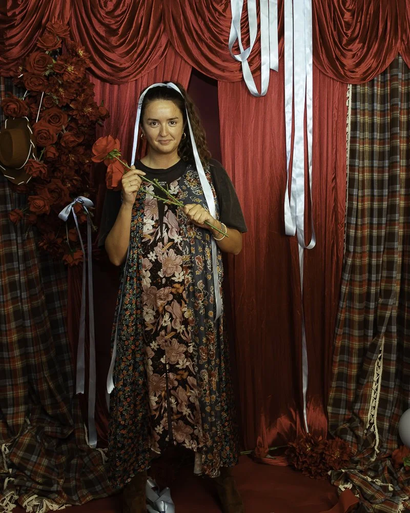 A woman standing in front of a red curtain backdrop, holding a red rose, surrounded by red roses and plaid curtains, dressed in a floral dress with a black overlay and brown boots.