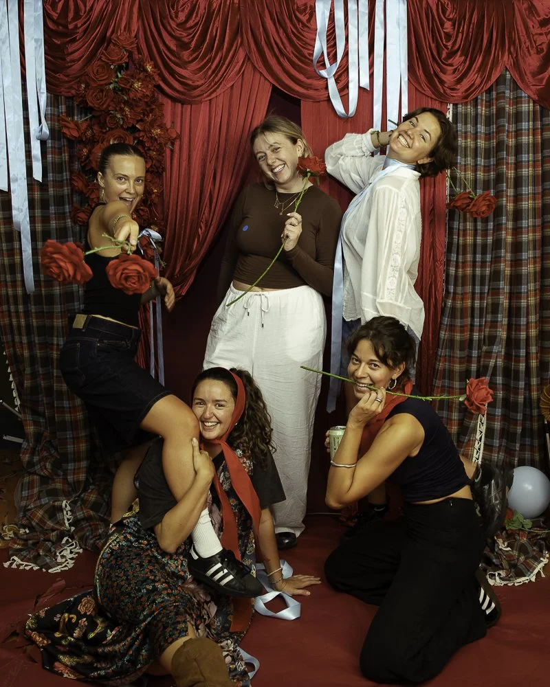 Six women are posing together in front of a red curtain with plaid and solid fabric drapes. They are smiling, holding roses, and dressed in casual and festive clothing, suggesting a celebratory or themed gathering.