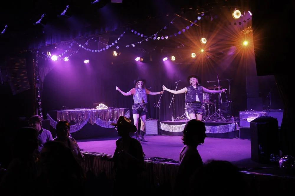Two performers on stage with guitars in hand, wearing cowboy hats and denim outfits, performing at a lively show with colorful stage lights, an audience in front, and various musical instruments on stage.