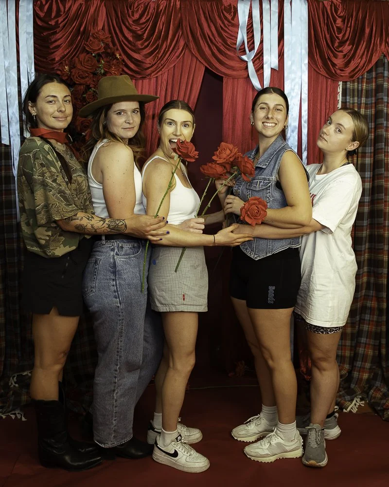 Five women standing in front of red curtains with blue ribbons, holding red roses, posing for a photo.