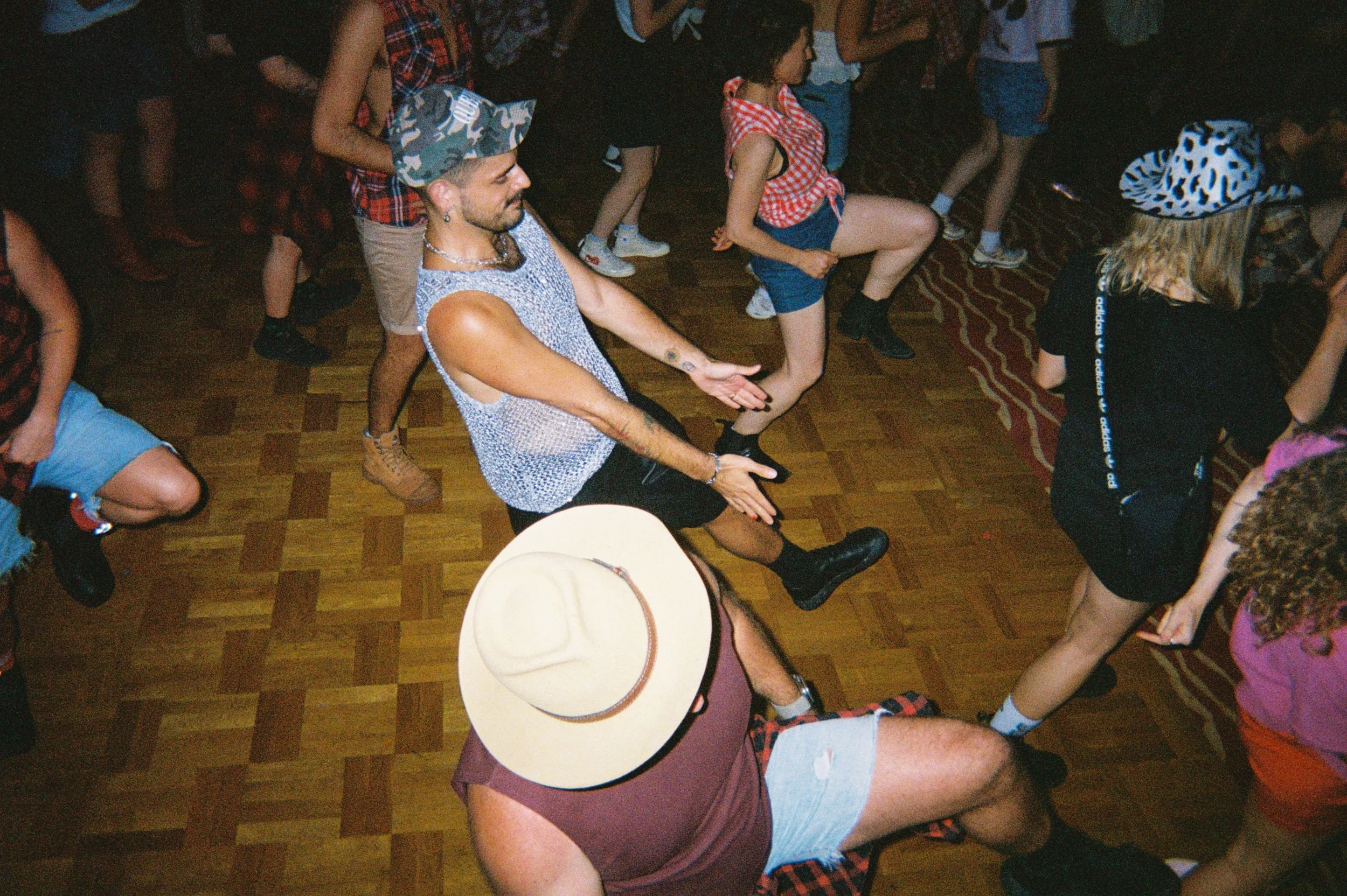 People dancing at a crowded party or dance event, with some individuals wearing casual clothing like tank tops, shorts, and hats, and others engaging in dance moves on a wooden floor.