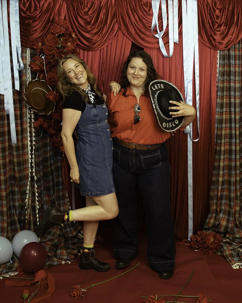 Two women posing in front of a red curtain with streamers, balloons, and red roses. One woman is wearing a denim dress, black t-shirt, and boots, while the other woman is dressed in a red shirt and holding a cowboy hat that reads 'Let's Disco.' They 