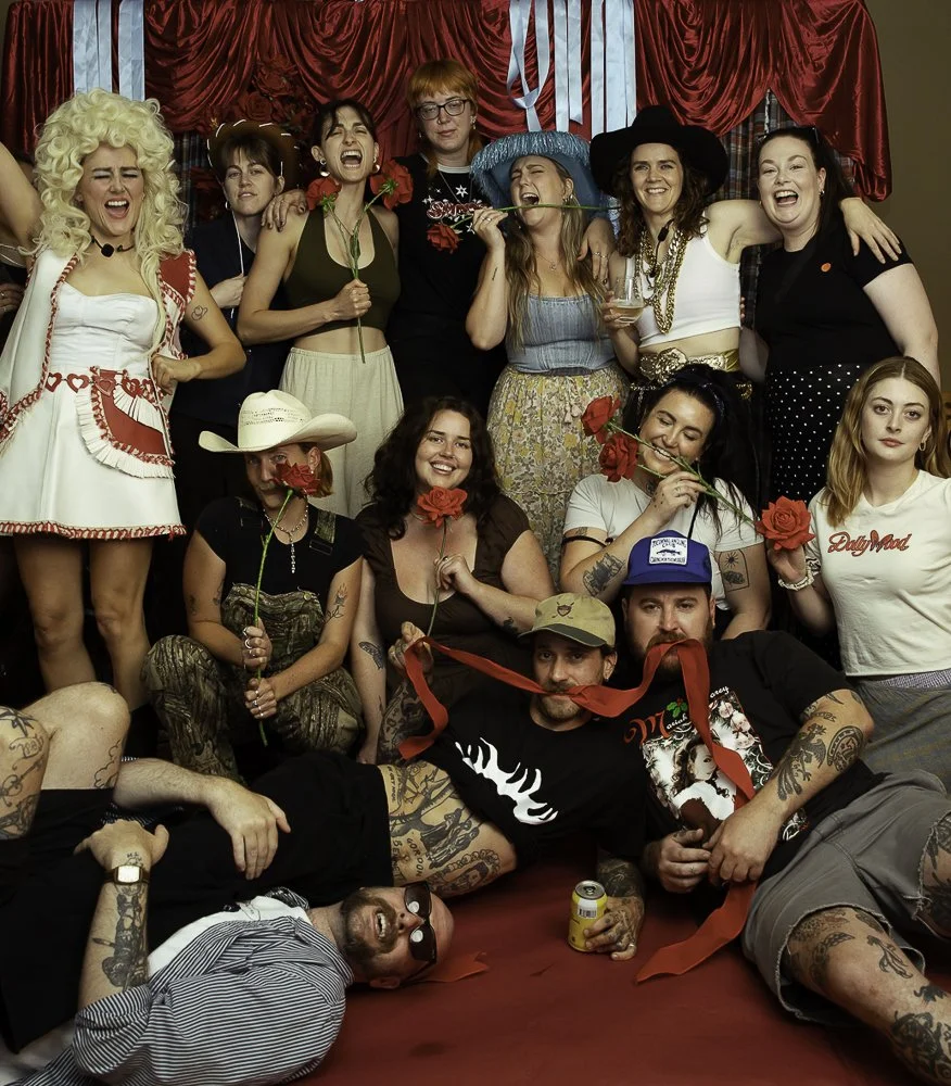 Group of women and men in costume at a celebration, some holding roses, in front of red curtains.