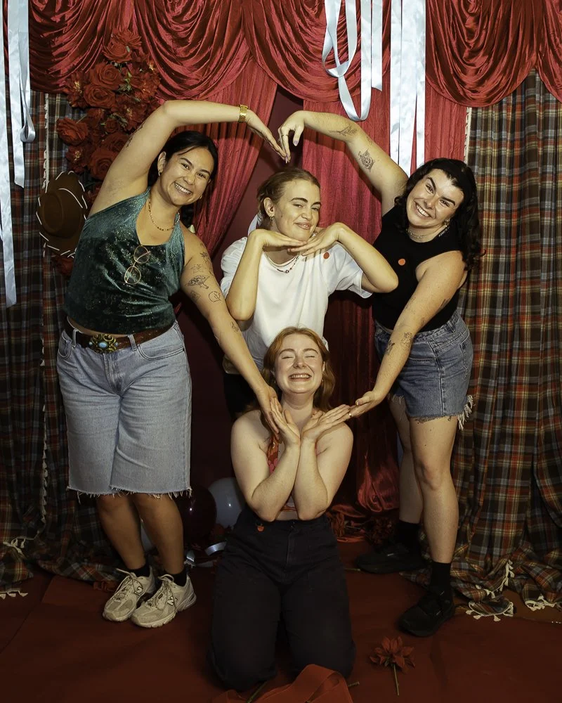Four women at a celebration posing together in front of a red curtain background, with two making a heart shape over the middle woman's head and another supporting her chin. All are smiling and appear happy.