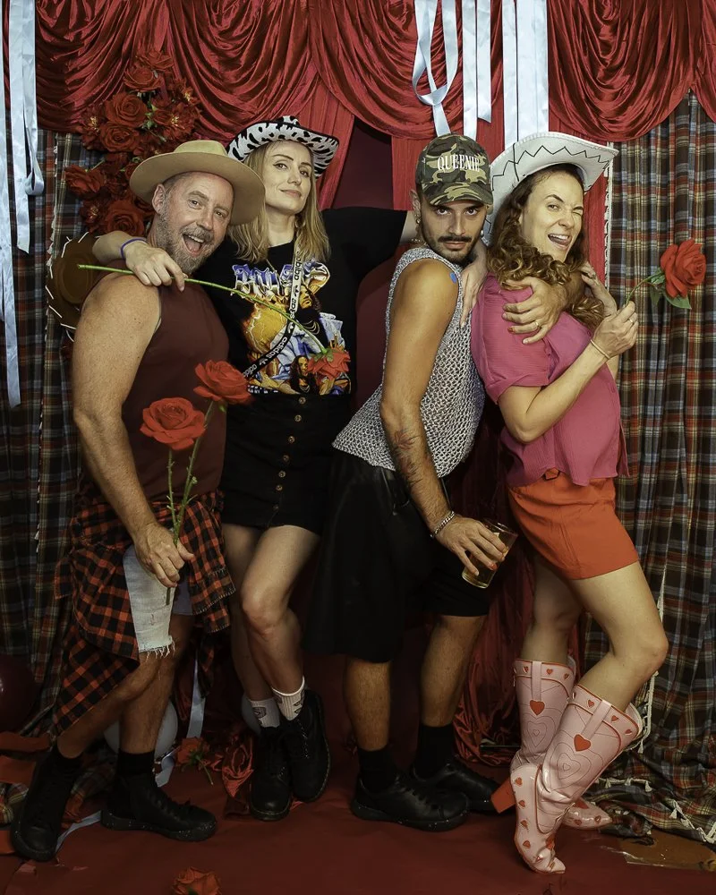 Four people dressed in colorful, playful costumes, holding roses and posing together in front of red and plaid curtains, suggesting a fun, party setting.