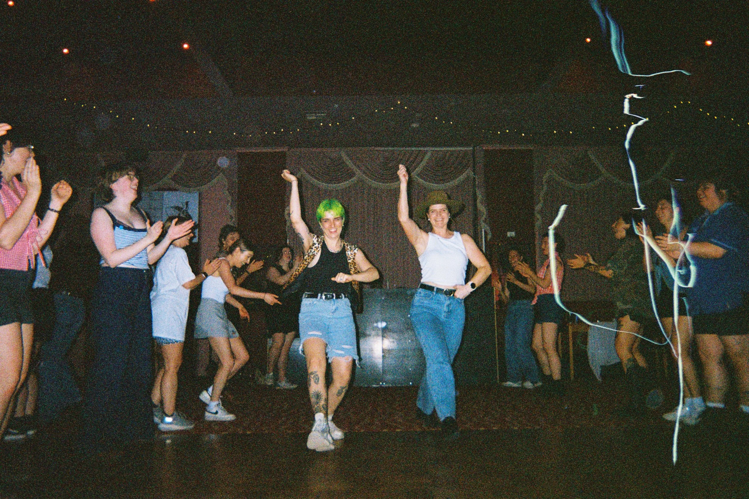 People dancing and enjoying themselves at a party or nightclub, with two women in the center dancing, one with green hair and the other with glasses, surrounded by others clapping and watching.