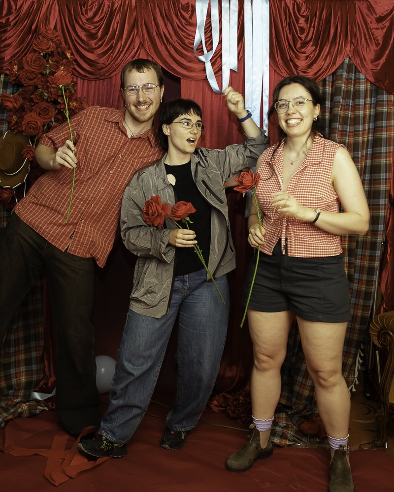 Three people standing in front of a red curtain backdrop, holding roses and smiling. Two women and one man, dressed casually, with decorative roses and ribbons in the background.