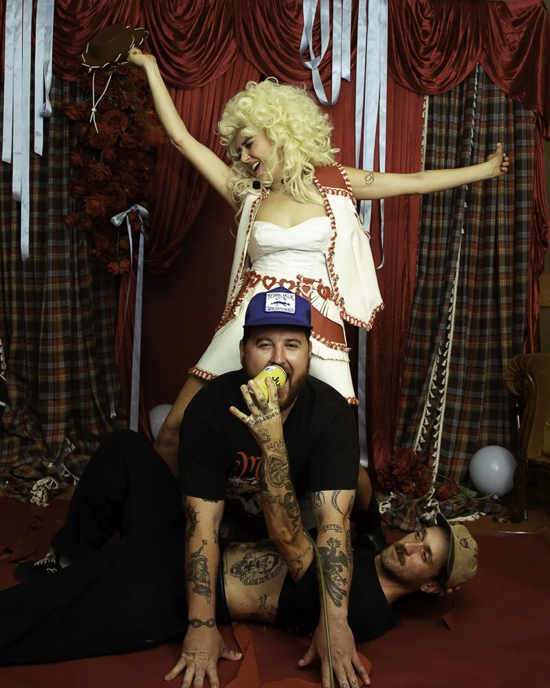 A woman in a white dress with red heart patterns and a blond curly wig stands with arms outstretched behind two men, one sitting and one lying on the floor. The man on the floor has tattoos, a tan cap, and is lying on his side, while the man in the m