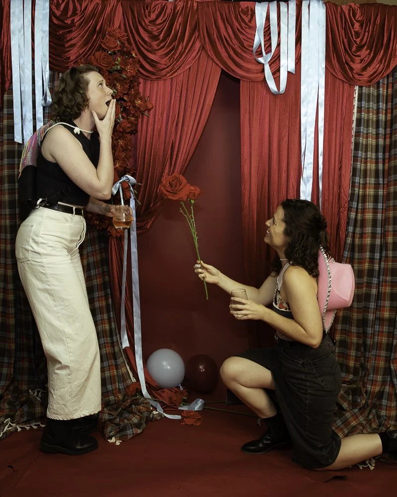 A woman on her knees offering a single red rose to another woman who is surprised and covering her mouth in shock at an indoor Valentine’s Day or romantic setting with red curtains, balloons, and romantic decorations.