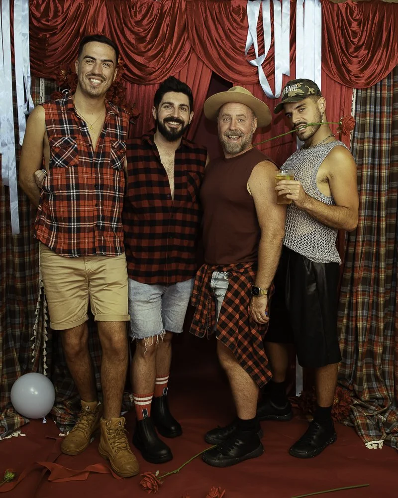 Four men standing in front of red curtains, dressed casually with some in plaid shirts, celebrating with roses and drinks.