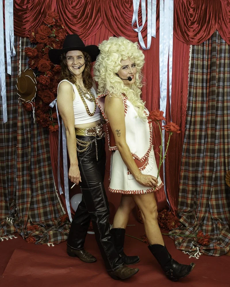 Two women dressed in costumes pose in front of a red floral and curtain backdrop, one with a black cowboy hat and leather pants, the other with a large curly blonde wig and holding red roses.