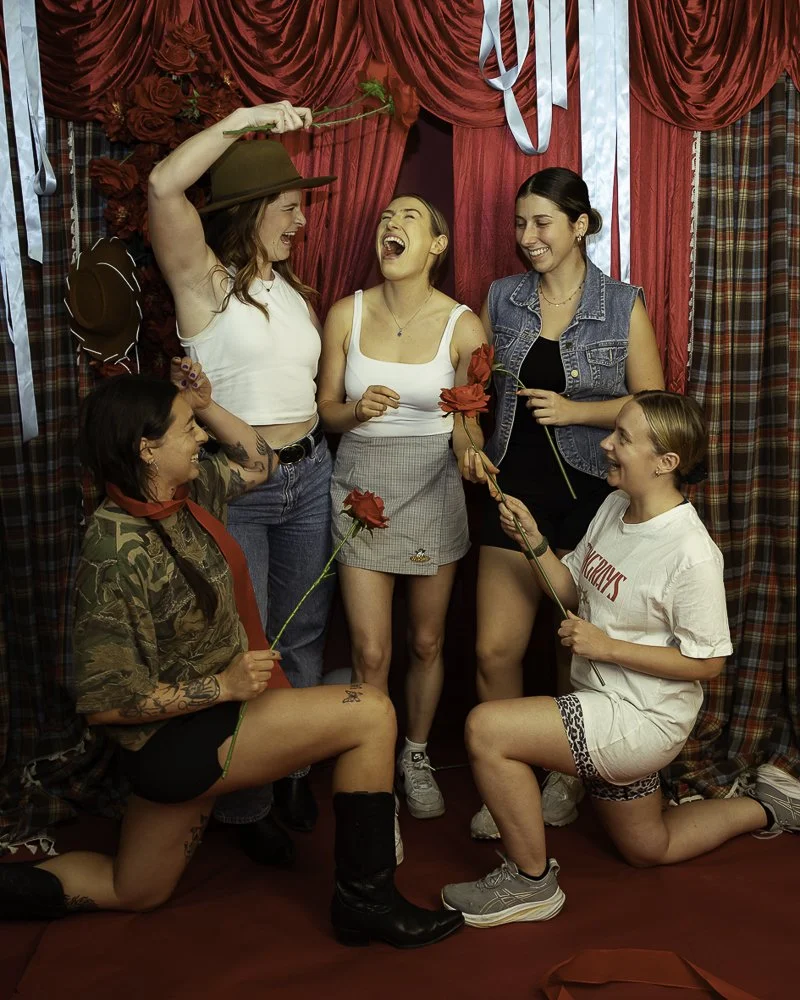 Five women and one girl celebrating in front of red curtains, with some holding roses and one wearing a cowboy hat, smiling and laughing together.