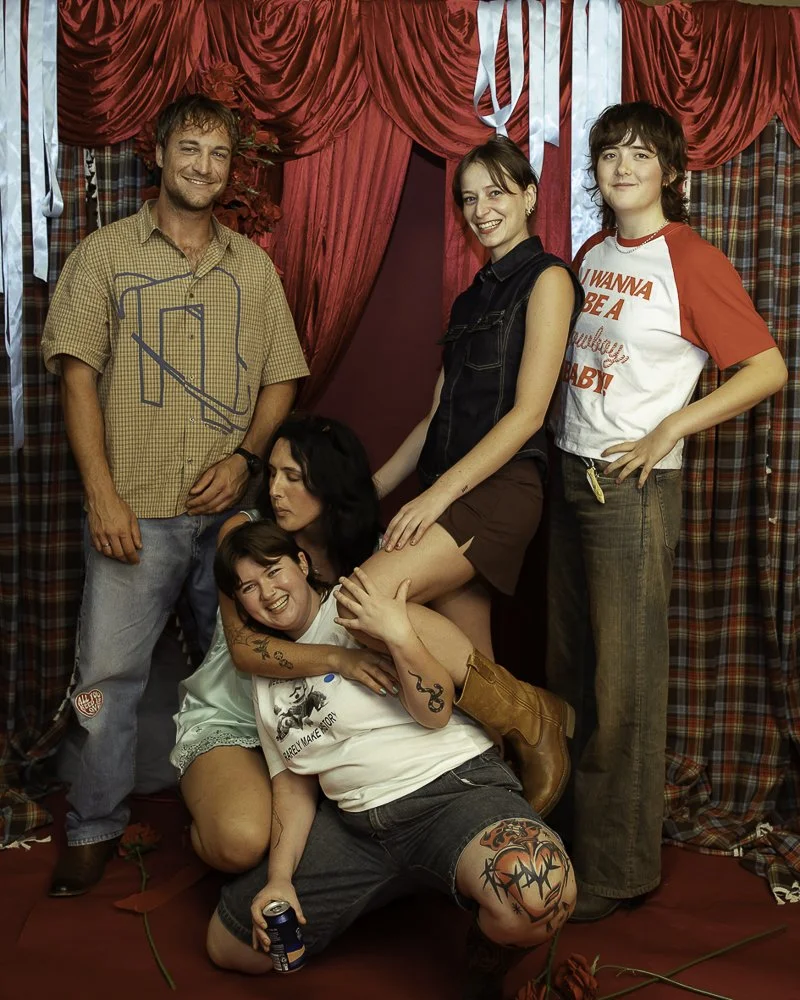 Group of five people posing in a room with red curtains, some smiling and one person sitting on the floor holding a drink, while two others are standing with hands on the sitting person's legs.