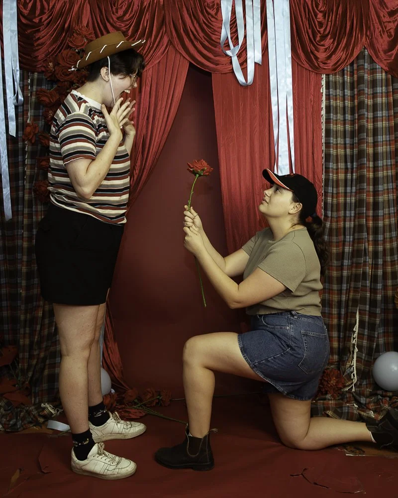 A man kneeling on one knee presenting a red flower to a woman with a surprised and happy expression, indoors with red curtains and plaid drapes.