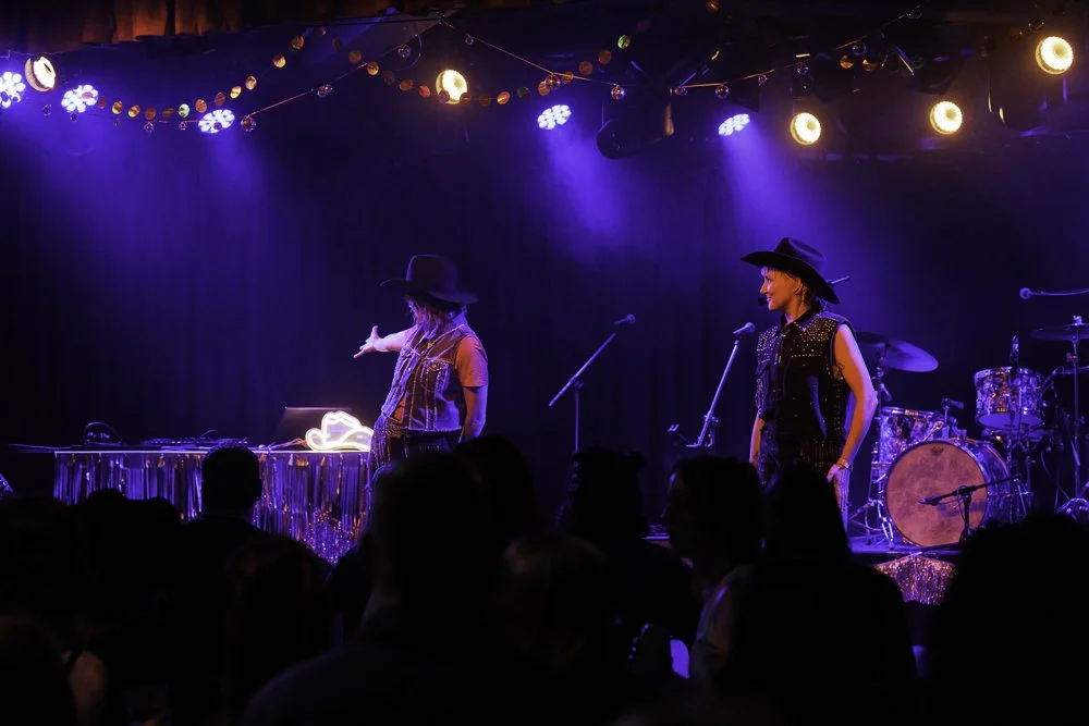 Two women wearing black hats perform on stage with musical instruments, colorful stage lights, and a dark background at a concert or musical event.