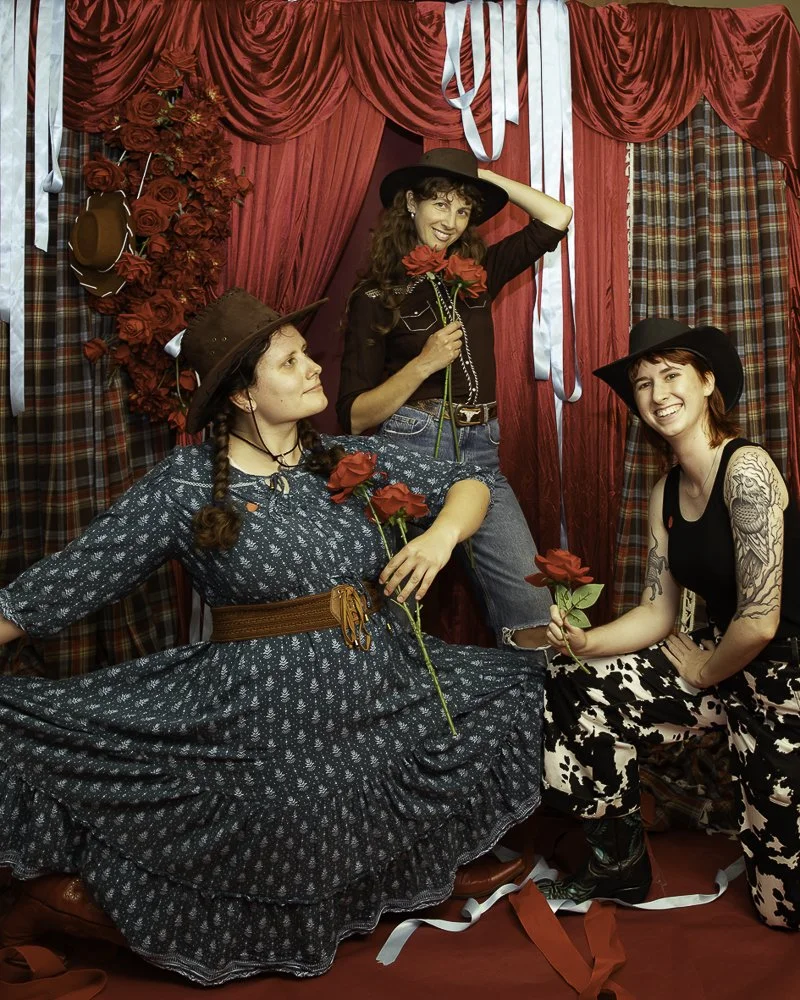 Three women dressed in vintage western attire inside a decorated red and plaid curtain backdrop, holding red roses and posing cheerfully.