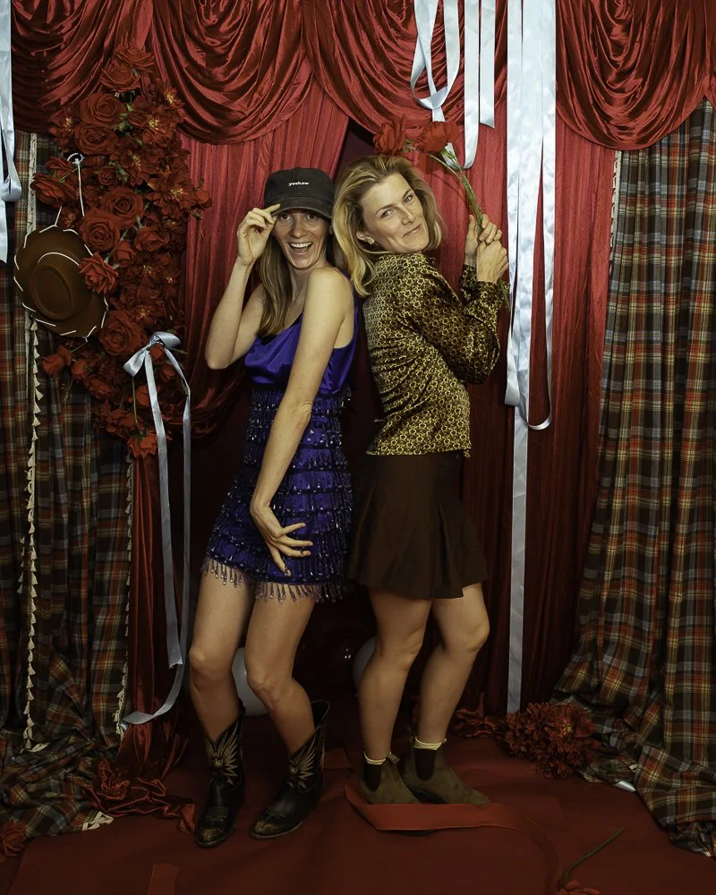 Two women posing in front of a red curtain backdrop, with roses and ribbons, one wearing a leopard print top and brown skirt, the other in a blue dress and cowboy boots, both smiling and holding roses.