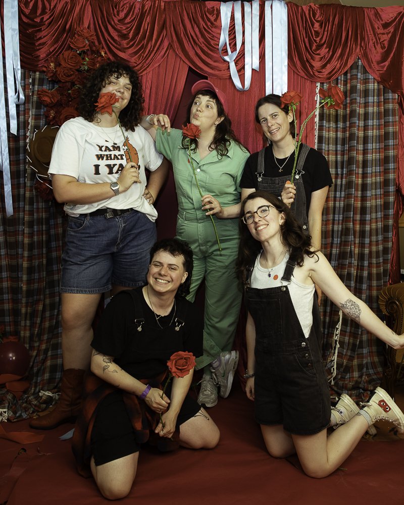 Five women posing with roses in a colorful, vintage-themed setting with red curtains and plaid drapes.