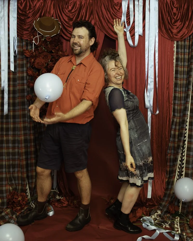 A man and woman standing back-to-back against a red curtain backdrop, celebrating at a festive event with balloons, roses, and streamers.