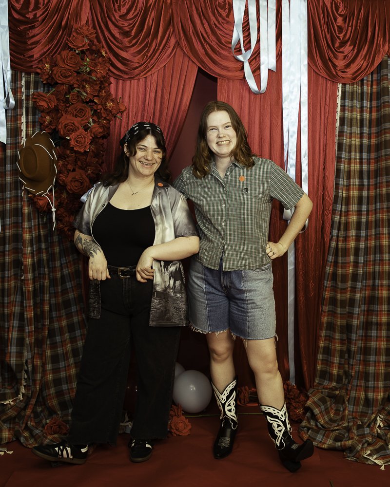 Two women smiling and posing in front of red velvet curtains with plaid and red floral decorations, balloons, and a cowboy hat.