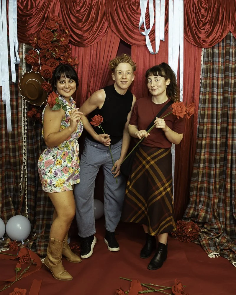Three women standing in front of a red curtain with roses and balloons, holding roses, in a decorated room.