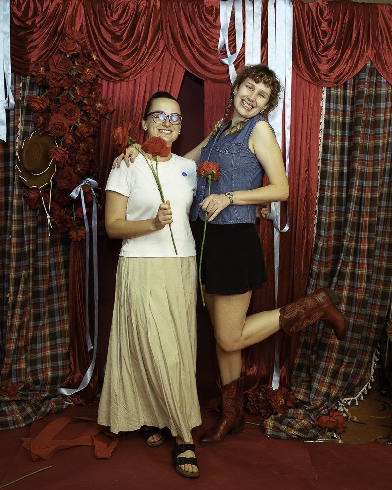 Two women smiling, holding red roses, standing in front of a red and plaid curtain backdrop with roses and ribbons