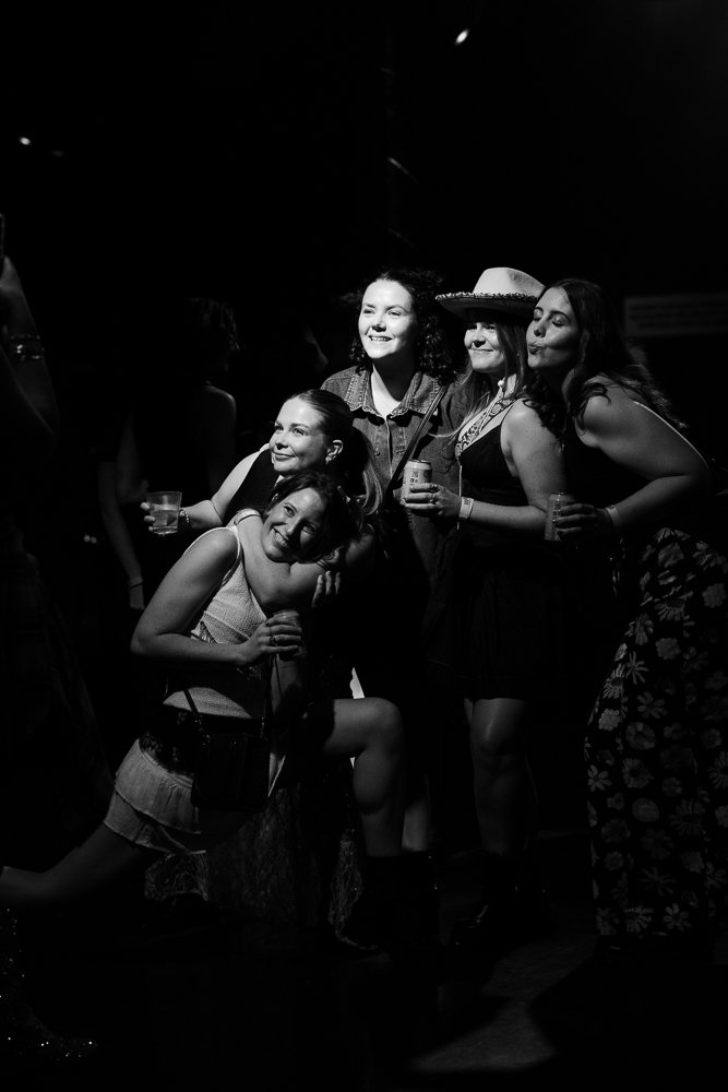 Group of five women at a social gathering, smiling and holding drinks in a dark setting.