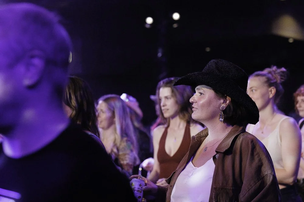 A group of women at a concert or event, with one woman in the foreground wearing a large black hat, brown jacket, white top, and earrings. The women are looking towards the stage or performer with interest. The background is dark with stage lighting.