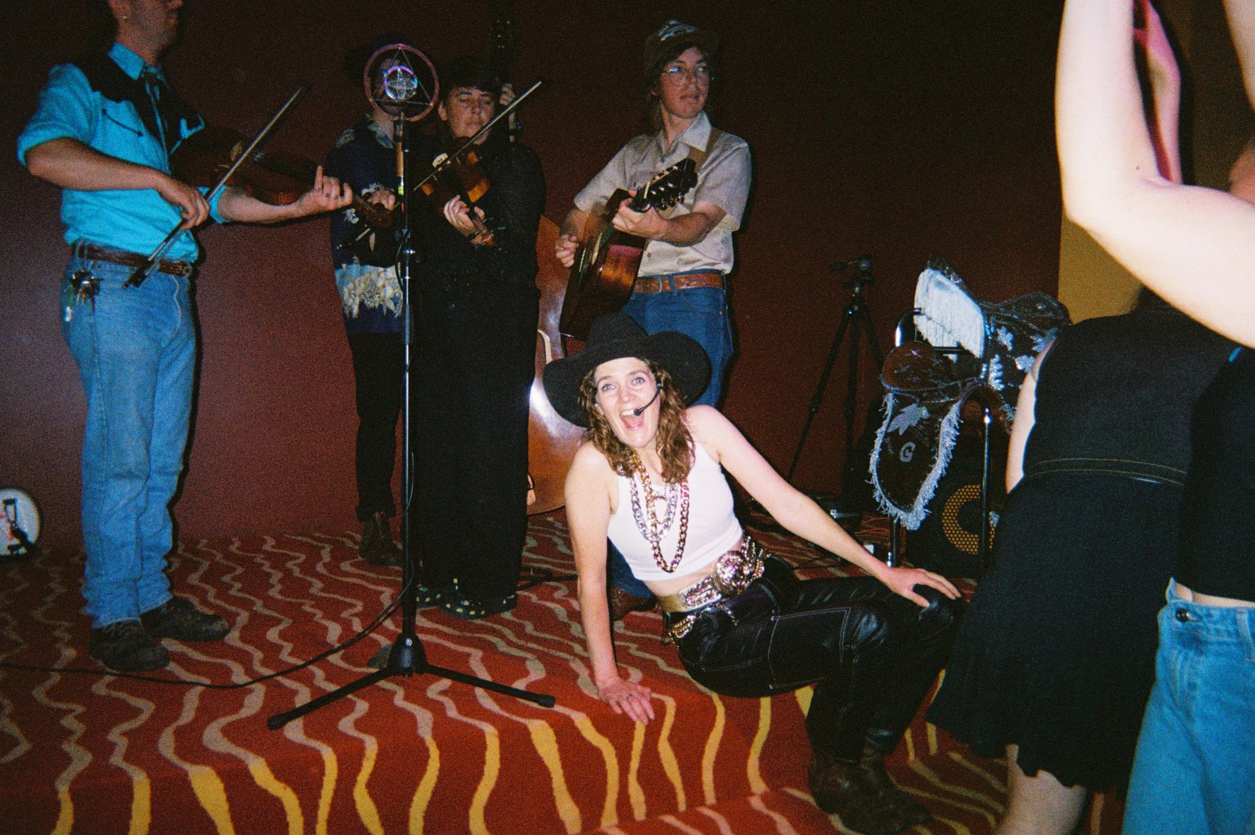 Group of people performing music on stage, with a woman in a witch costume sitting on the floor smiling.