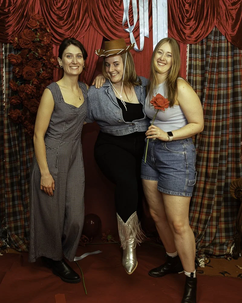 Three women posing in front of red and plaid curtains, with roses and ribbons, celebrating Valentine's Day. One woman is wearing a cowboy hat and metallic cowboy boots.