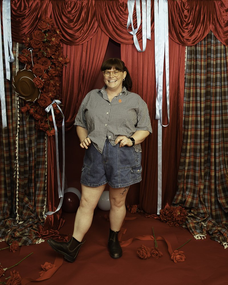 Woman smiling, standing in front of a red curtain backdrop decorated with roses, plaid curtains, balloons, and roses on the floor.