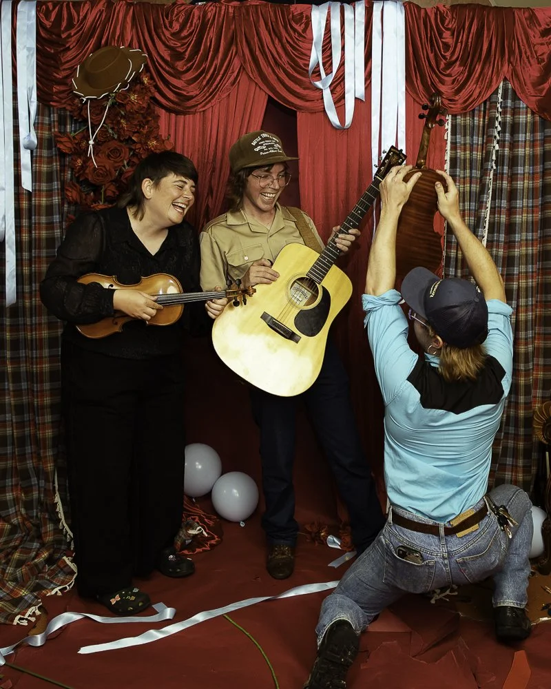 Three people are on a stage with red curtains, two women are playing guitars, one woman is wearing a black shirt and the other is wearing a beige shirt and a hat. A man with a blue shirt and a cap is kneeling and taking a picture of them with his cam