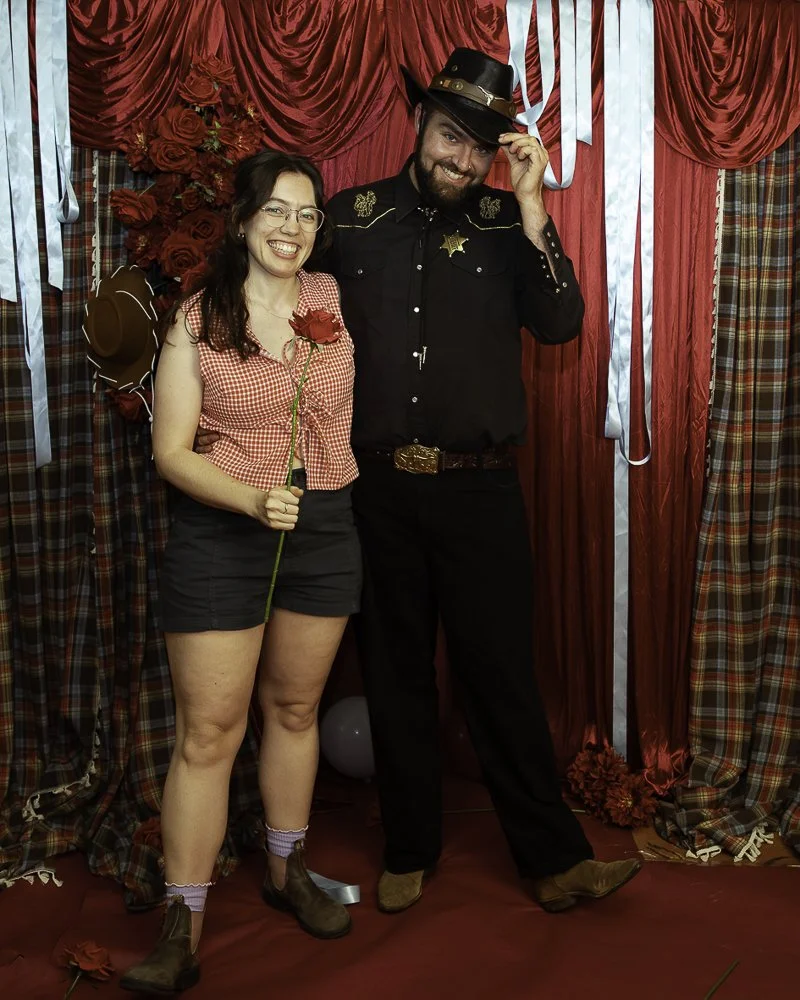 A woman and man dressed in costume, standing in front of red curtains with plaid accents, smiling and posing for the photo. The woman holds a red rose, wearing glasses, a red checkered shirt, shorts, and boots. The man is dressed as a sheriff with a 