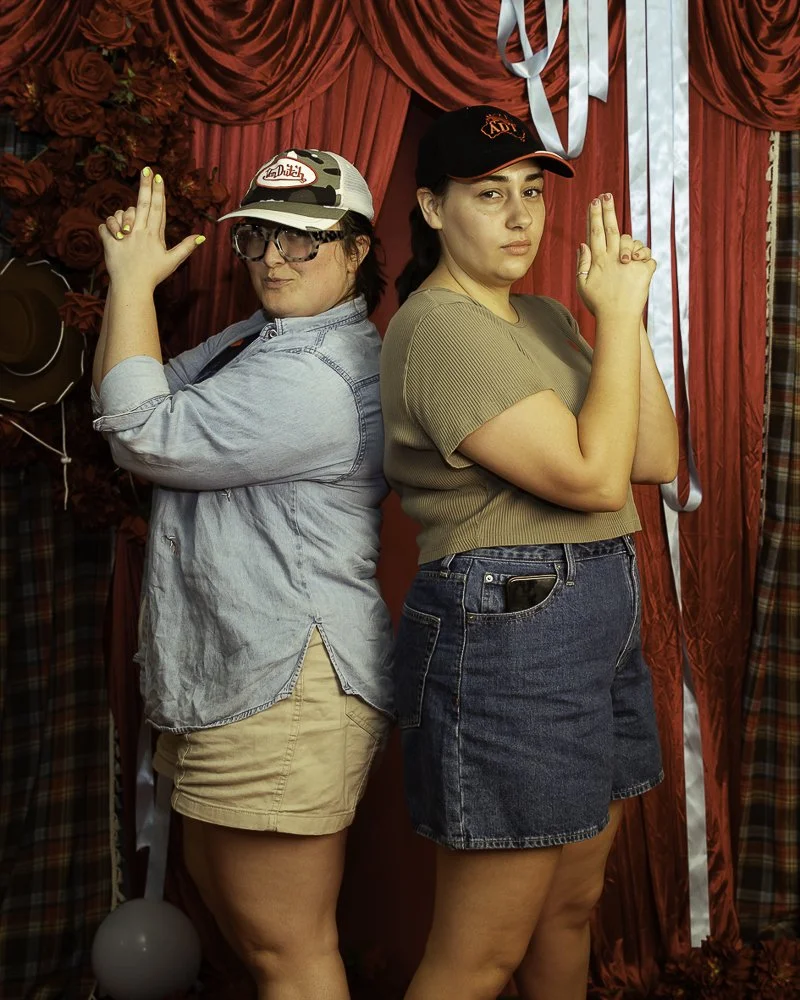 Two women standing back to back in front of red curtains and floral backdrop, making peace signs with their hands.