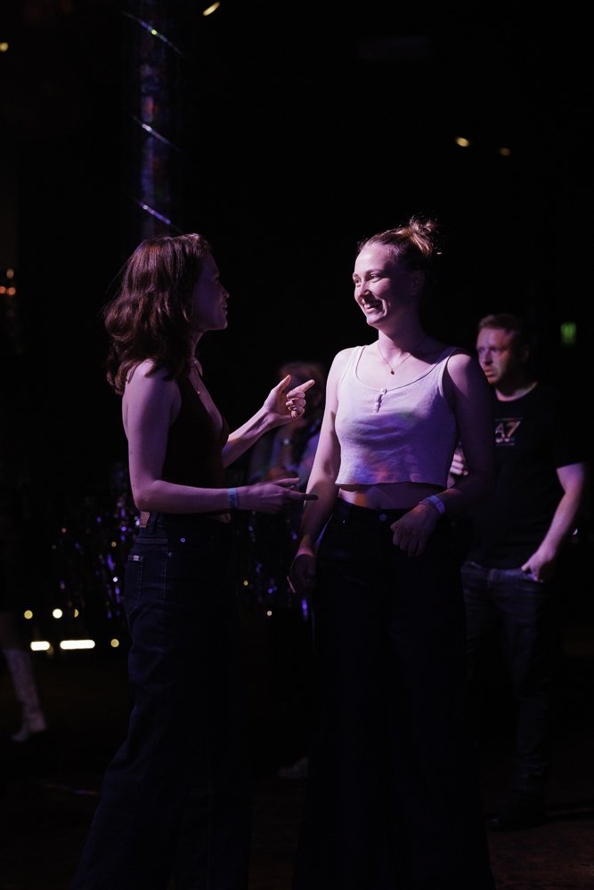 Two women are engaged in a conversation at a dark, indoor event with colorful lighting. The woman on the left has shoulder-length wavy hair and is wearing a sleeveless top and high-waisted jeans. The woman on the right has her hair in a bun and is we
