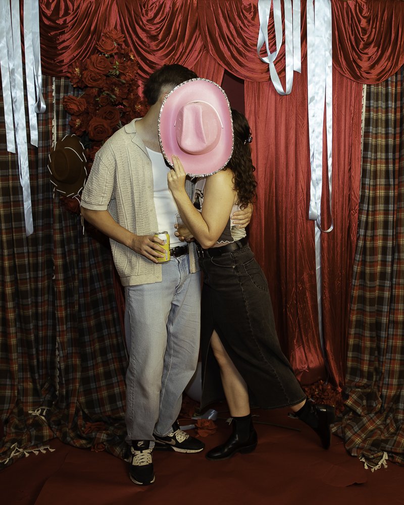A couple standing in front of red and plaid curtains, with roses in background. The woman is holding a pink sombrero mask in front of her face, and the man is holding a drink, with their arms around each other.