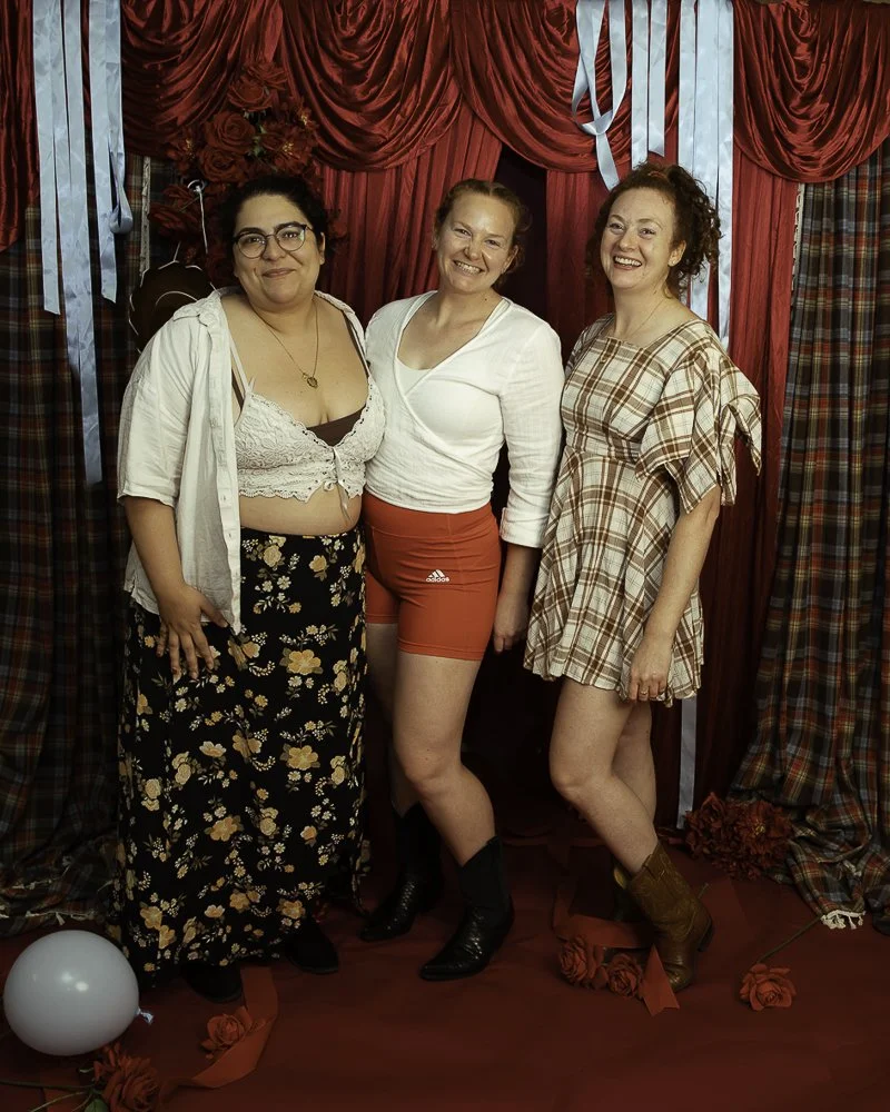 Three women smiling and posing together in front of a red curtain with ribbon and floral decorations, wearing casual and vintage-inspired outfits.