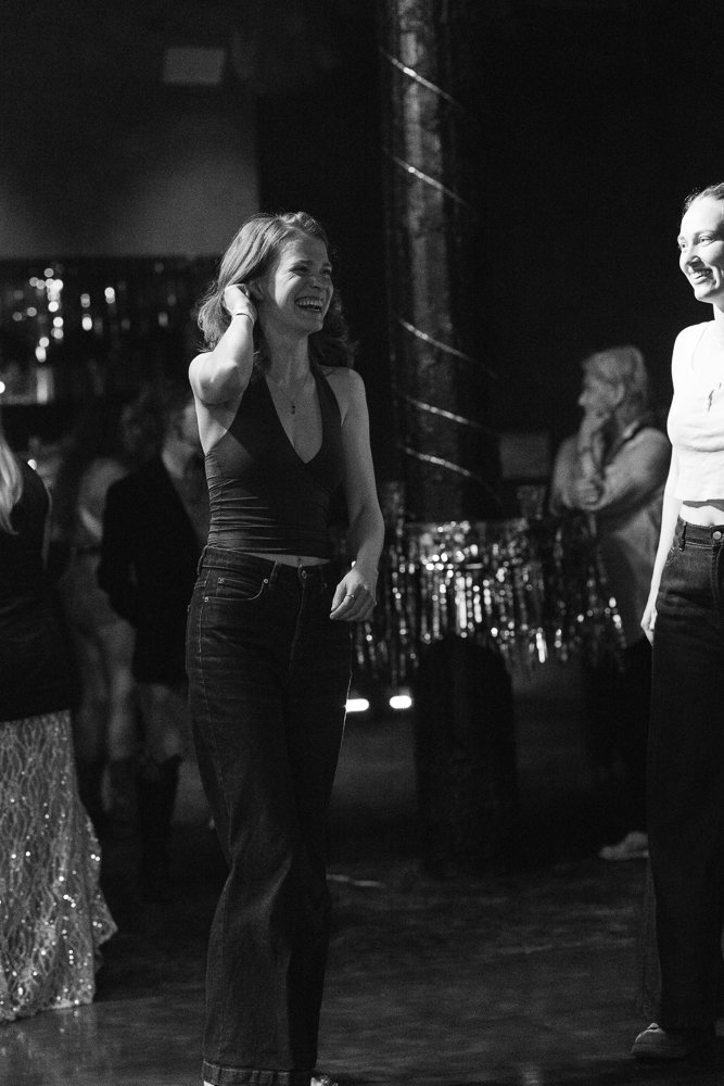 Two women smiling and enjoying themselves at a social gathering or party, standing near a decorated background with other people in the background.