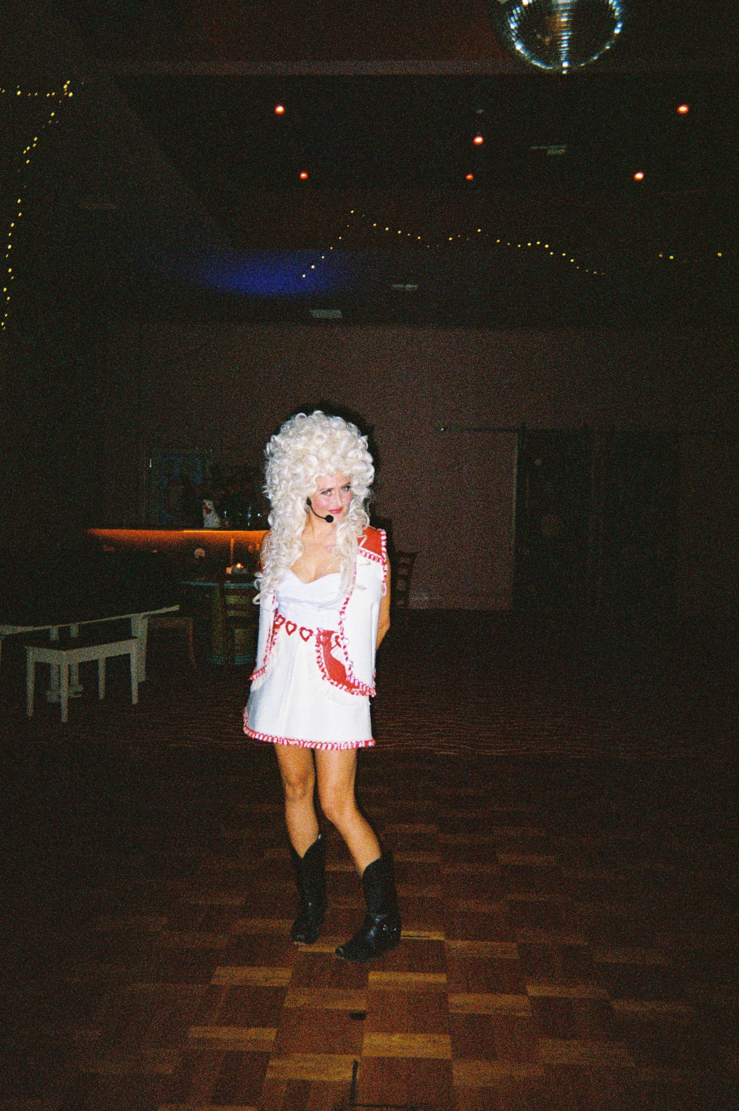 A woman in a white dress with red heart and lipstick patterns, black boots, and a large curly white wig standing on a wooden dance floor in a dimly lit room, possibly at a party or costume event.
