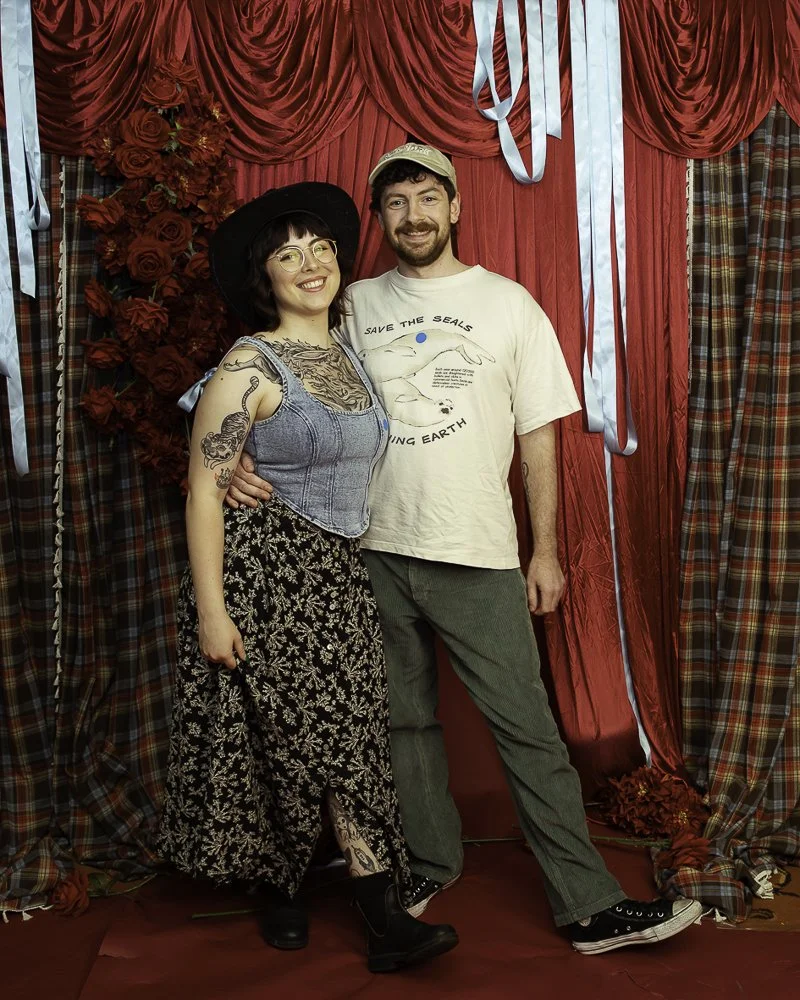 A smiling couple standing in front of a red and plaid curtain backdrop, with the woman wearing a black hat, glasses, a denim top, and floral wide-leg pants, and the man wearing a beige baseball cap, a graphic t-shirt, and green pants.