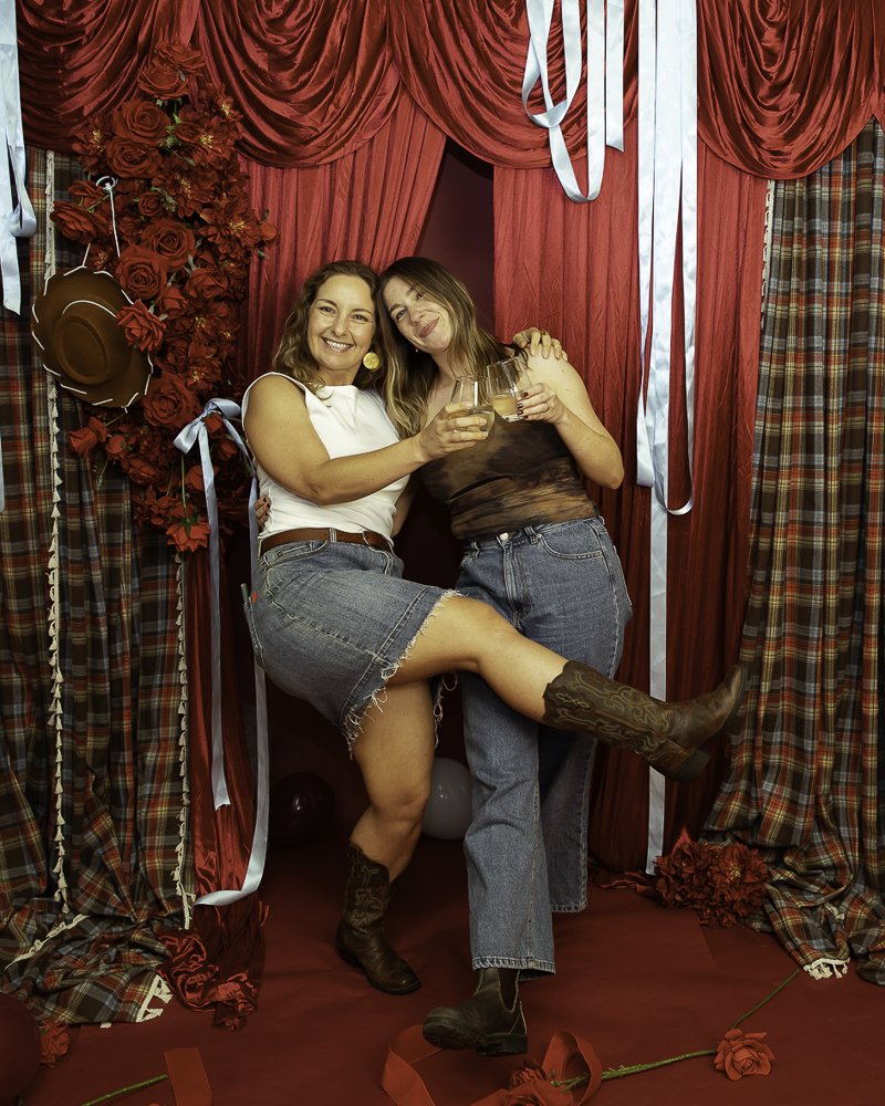 Two women celebrating in front of red and plaid curtains, holding glasses of wine, with one woman wearing cowboy boots and the other wrapping her arms around her, smiling.