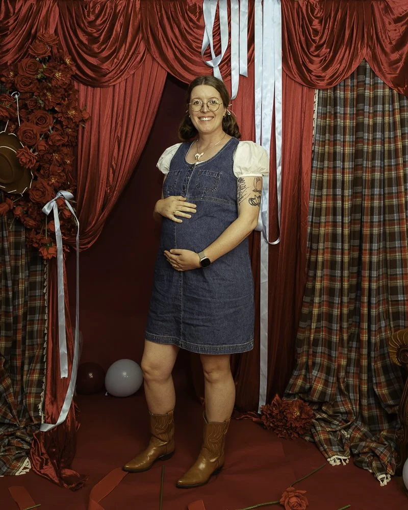 A woman with glasses, wearing a denim dress over a white puffed-sleeve shirt, standing in front of red and tartan curtains with flowers and balloons around, smiling and holding her pregnant belly.