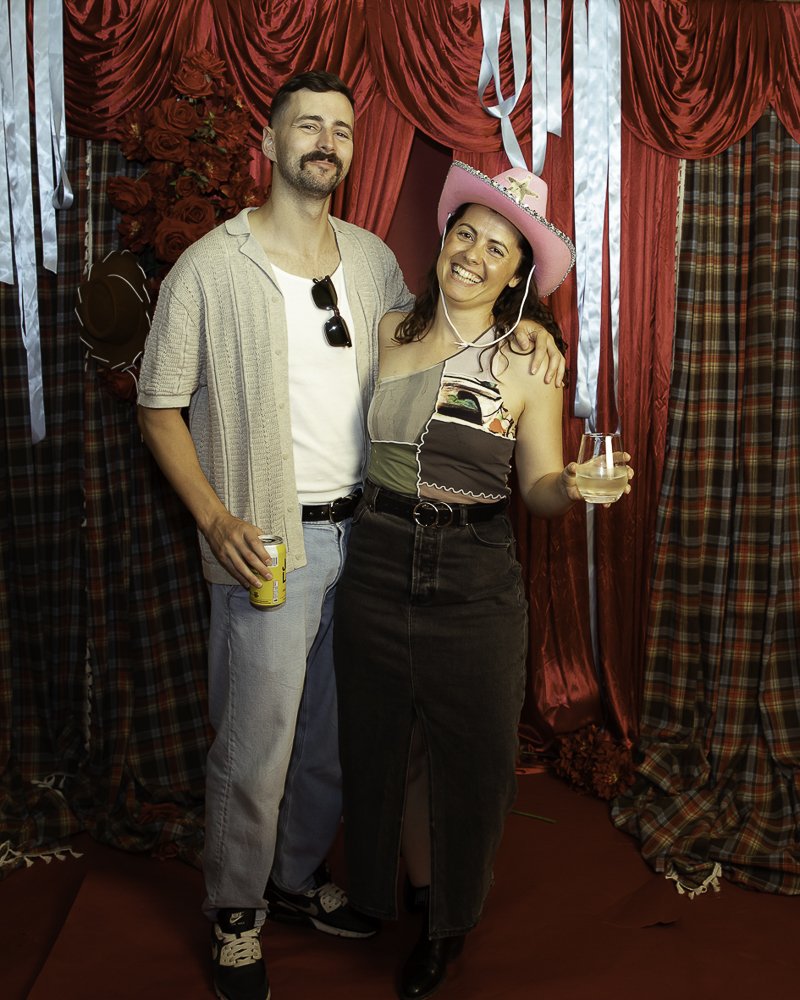 A man and woman posing together at a party, the woman wearing a pink cowboy hat and holding a glass of wine, with red and plaid curtains in the background.