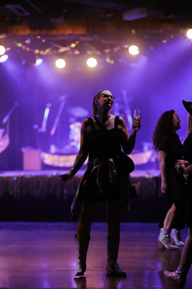 A woman in glasses holding a drink in a dimly lit dance venue with purple stage lighting and string lights, with other people in the background.