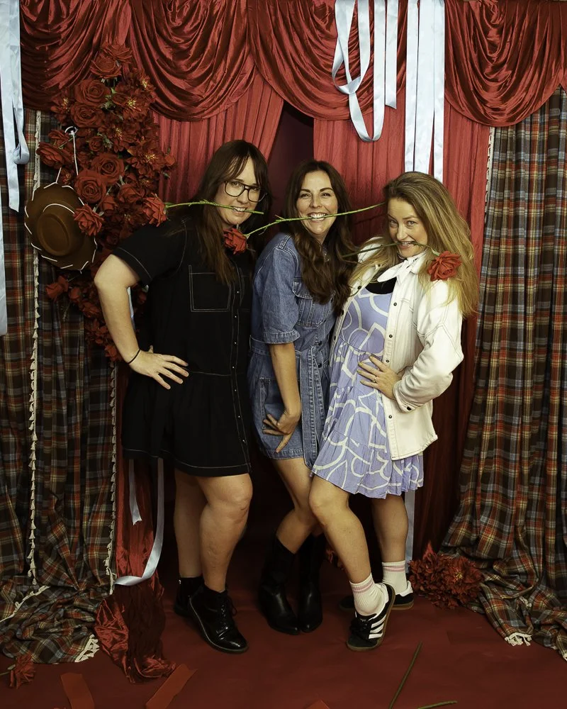 Three women standing in front of red curtains, holding roses in their mouths, with floral and plaid decorations around them.