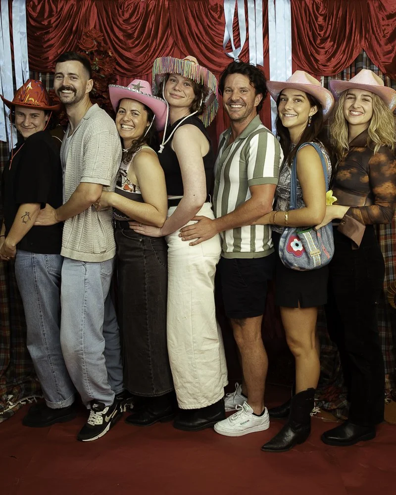 Group of seven friends at a party, wearing colorful hats and standing in front of a red curtain with blue streamers.