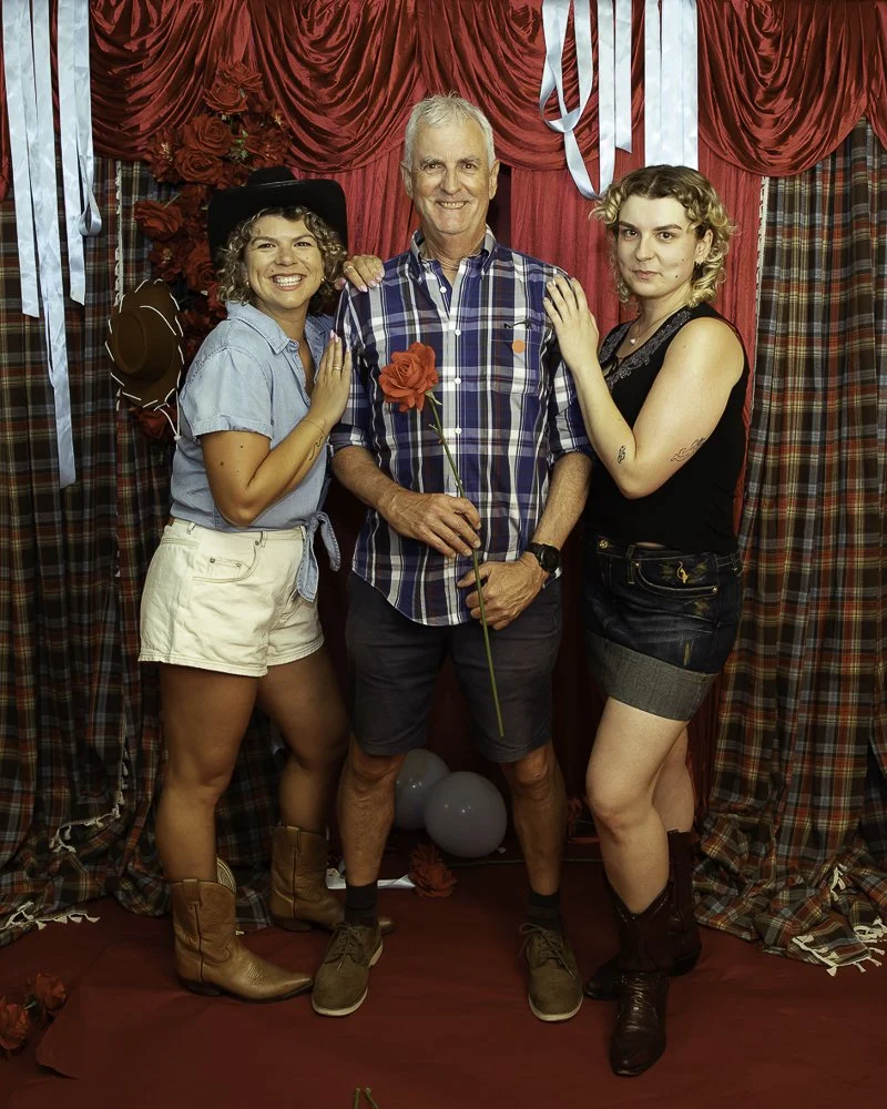 Three people standing together at a decorated event, with pink curtains, red flowers, balloons, and plaid drapes in the background. The man in the center holds a red rose and wears a plaid shirt, while the women on each side wear casual outfits and c