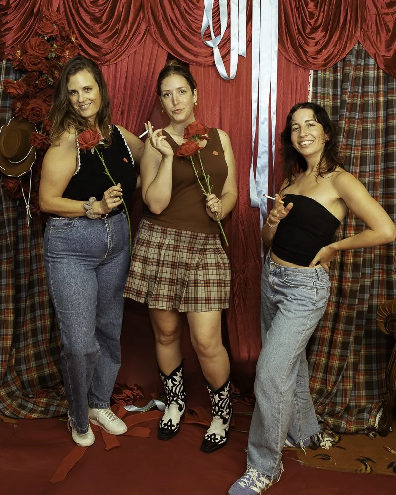 Three women standing in front of a red curtain with roses and plaid fabric, holding roses and cigarettes, dressed casually with a lively atmosphere.