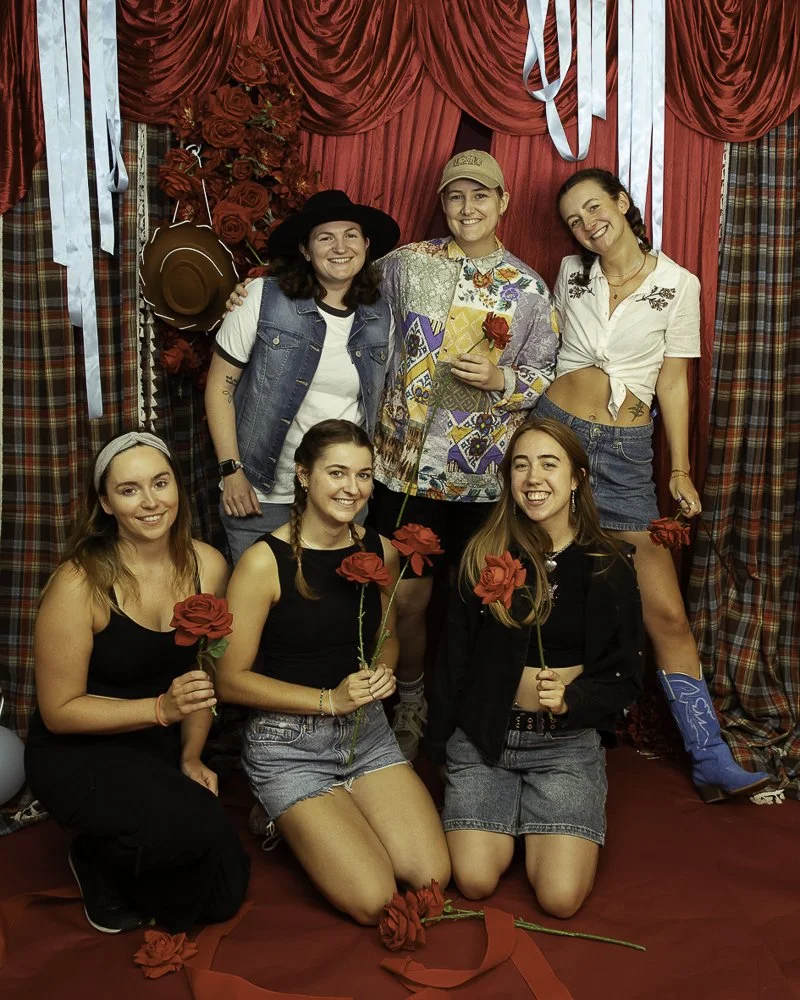 Six women posing in front of a red curtain backdrop decorated with roses and streamers; some are holding roses, and one is wearing cowboy boots and a hat.